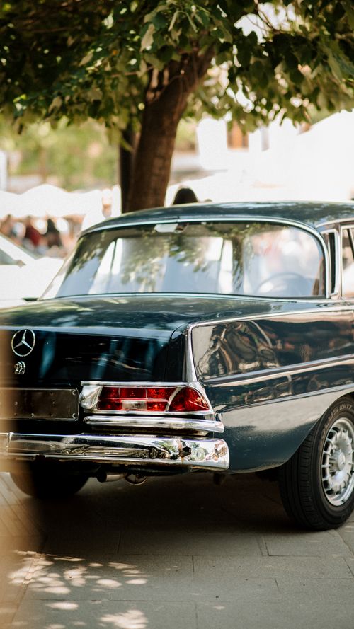 Free Vintage Mercedes-Benz car under a tree, captured in a sunny street in Kosovo, showcasing elegant retro design. Stock Photo