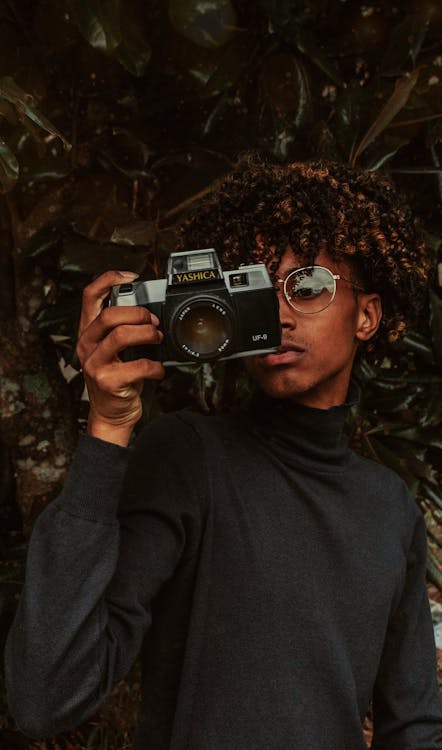 Free Stylish African American young male millennial in turtleneck and eyeglasses taking picture on vintage photo camera while standing near green tree in park Stock Photo