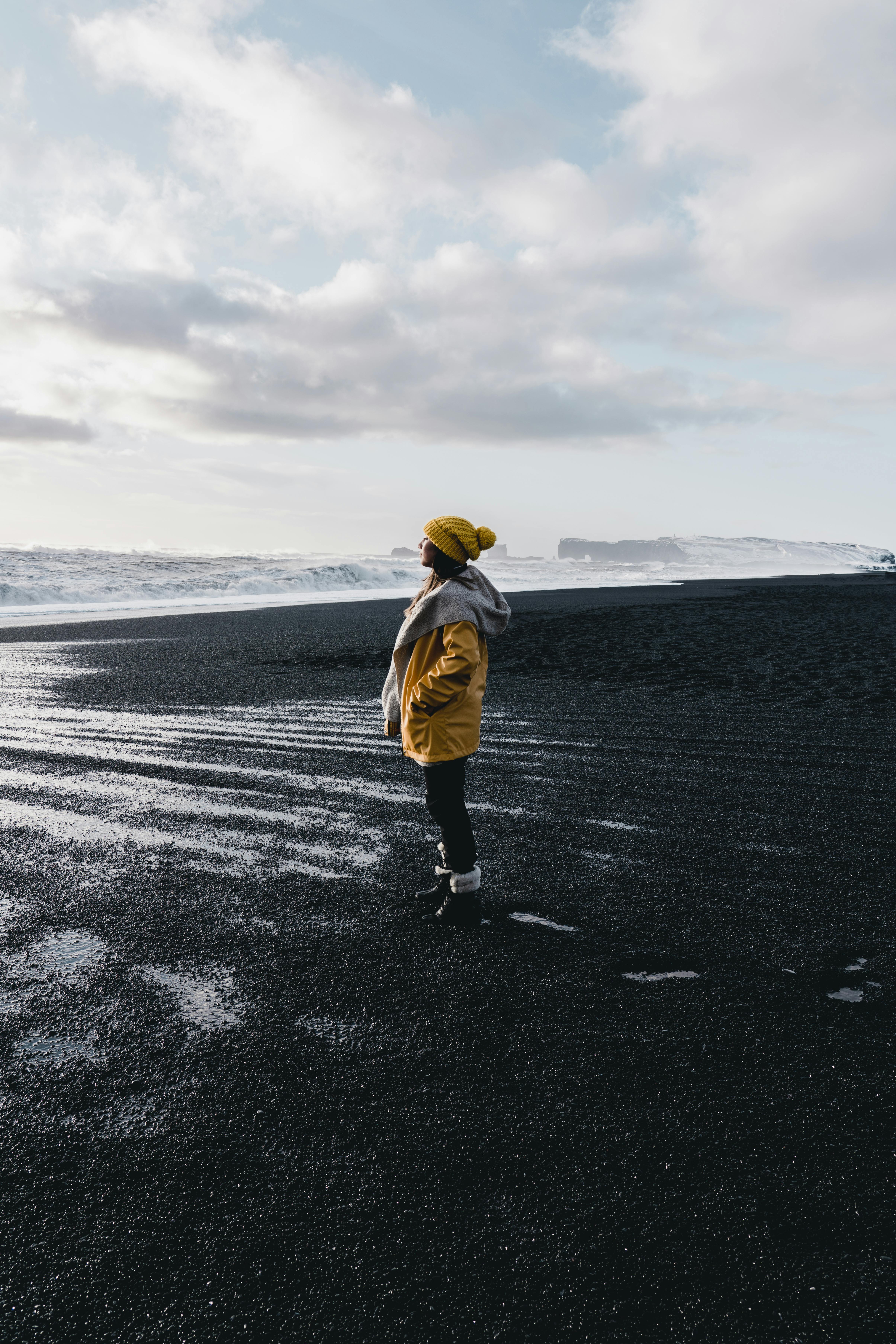 A person in a yellow jacket stands on a black sand beach in Vik, Iceland, under a cloudy sky.