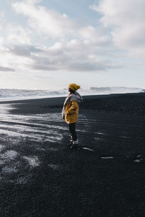 Free A person in a yellow jacket stands on a black sand beach in Vik, Iceland, under a cloudy sky. Stock Photo