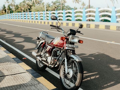 Free A vintage motorcycle parked on a sunlit road in Cherthala, Kerala, with a classic bridge in the background. Stock Photo
