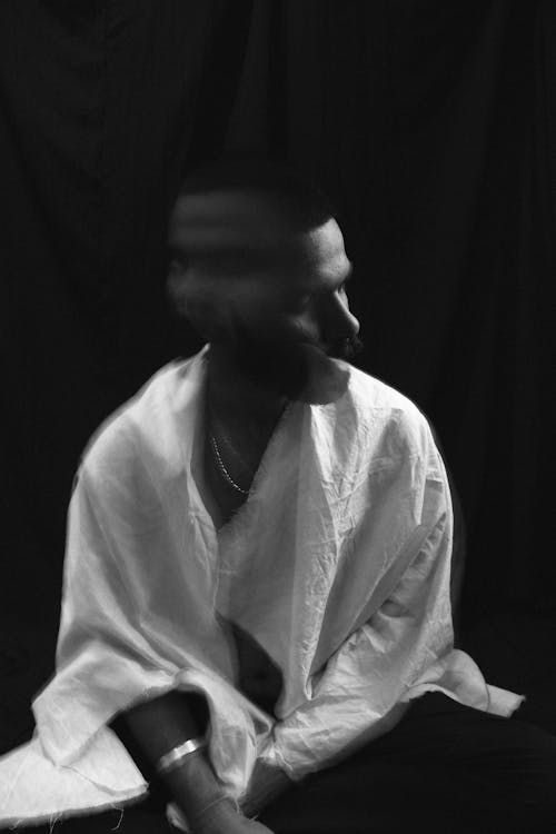 Black and white of lonely melancholic male in white shirt shaking head in motion sitting against black background