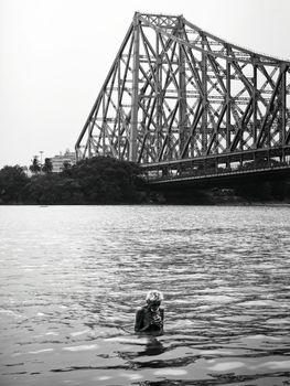 Black and white photo of a man bathing in a river beneath a steel truss bridge.
