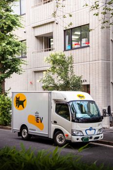 A compact electric delivery truck is parked on a street in Tokyo, Japan, with modern architecture in the background.