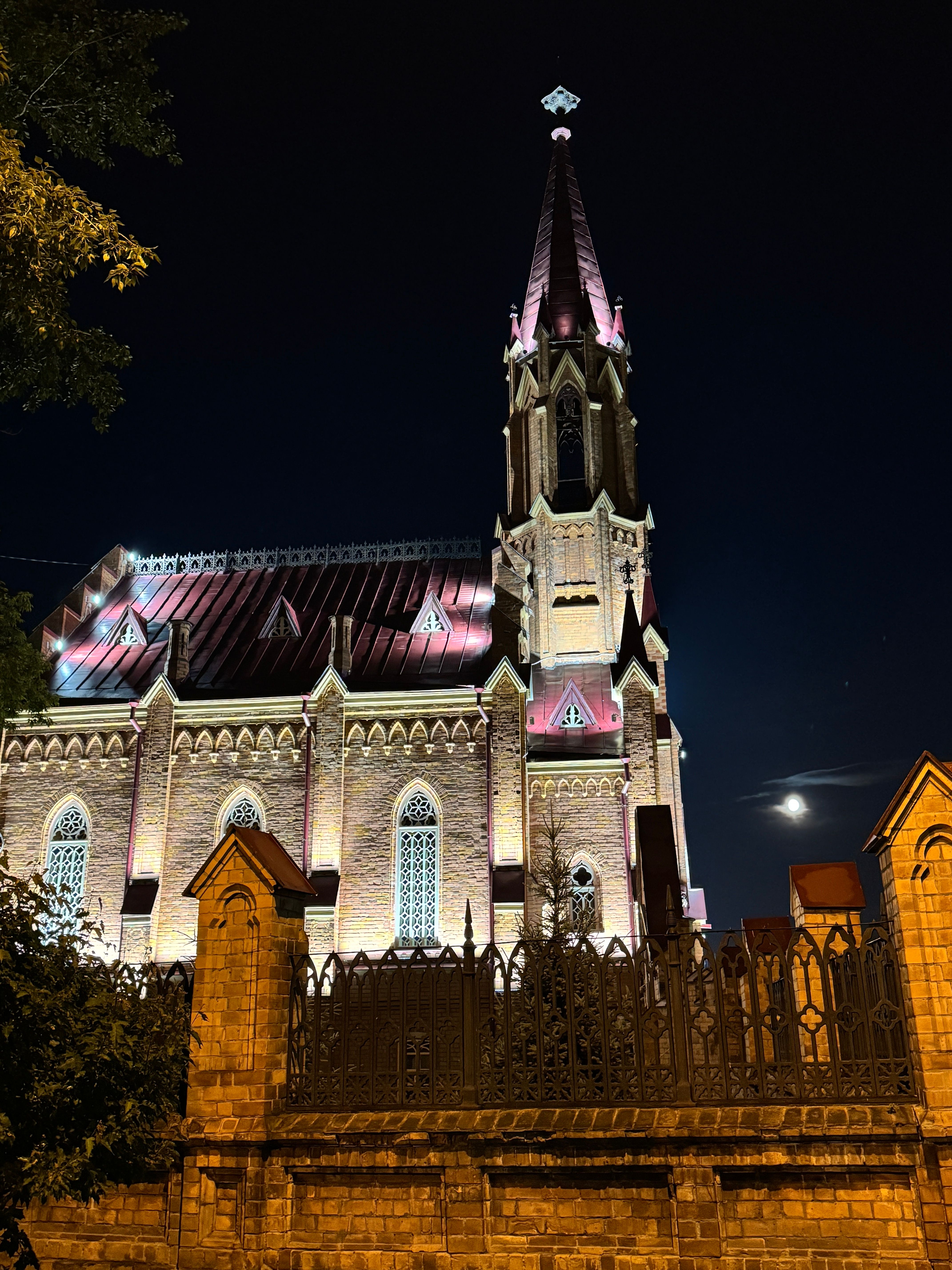 Dramatic night shot of a Gothic-style cathedral with a lit spire and moonlight accentuating its architectural features.