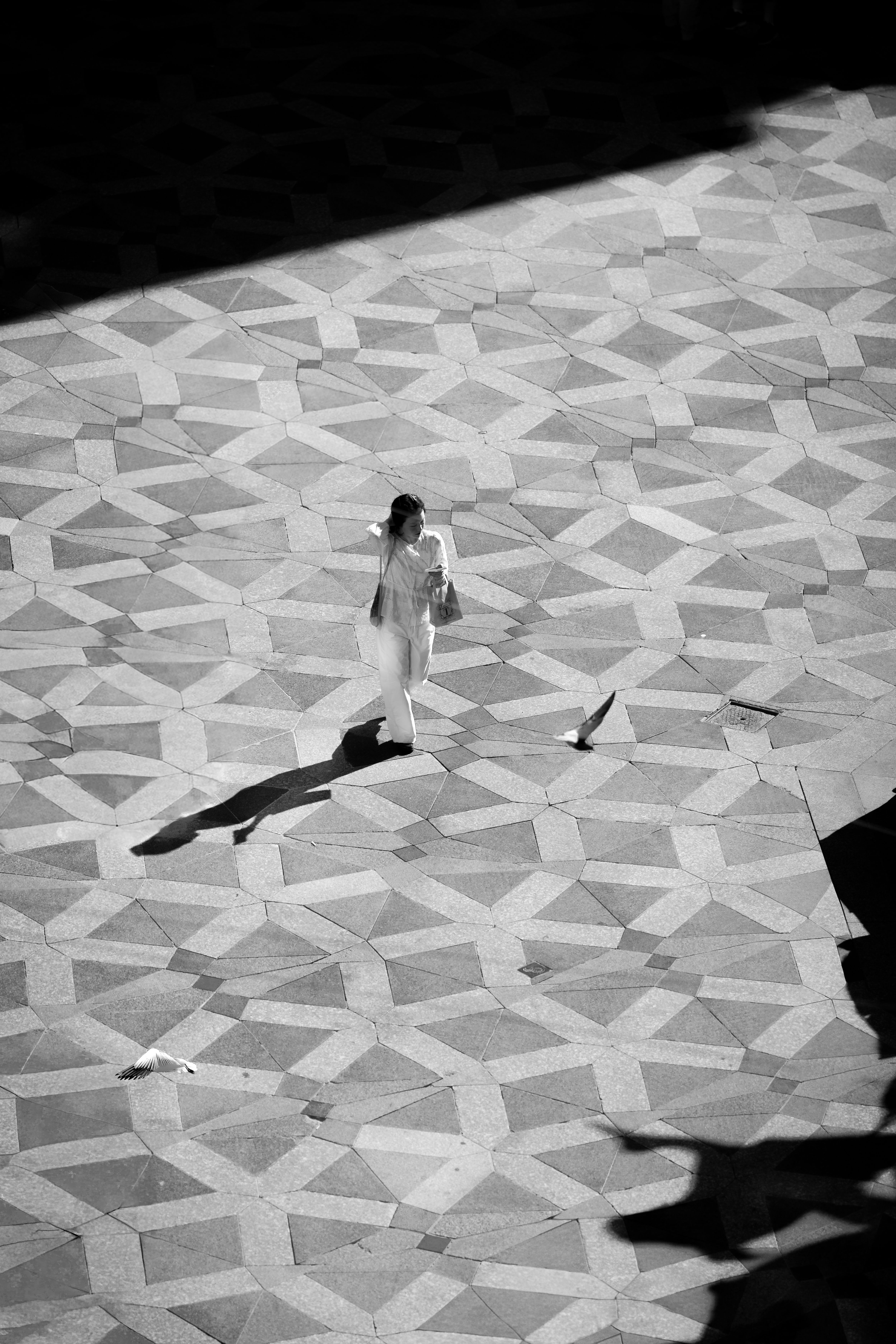 Black and white aerial photo of a woman walking on geometric patterned pavement with shadows.
