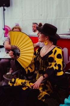 Elegant woman in a polka dot dress holds a fan while sitting. Captures Hispanic cultural attire.