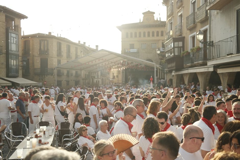Festival multitudinario en la plaza de un pueblo histórico europeo, con gente vestida de blanco disfrutando del buen tiempo.