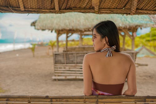Back view of confident young ethnic female in swimsuit and sunglasses leaning on beach cafe bamboo furniture and looking over shoulder during summer holidays in tropical resort