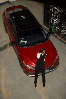 A fashionable young woman poses on a red car in an urban environment, viewed from above.