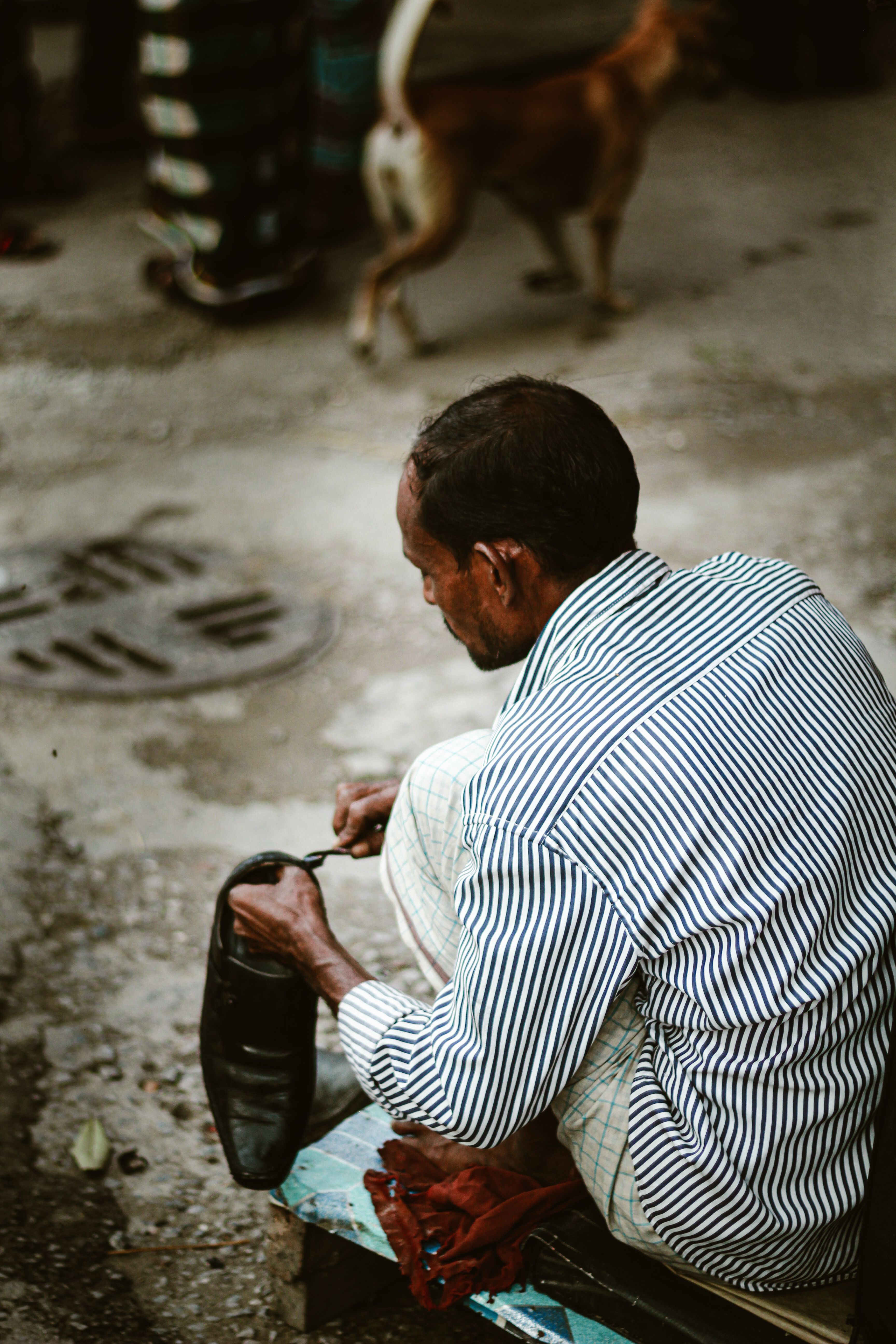 An adult shoe shiner polishes footwear outdoors in Dhaka, Bangladesh.