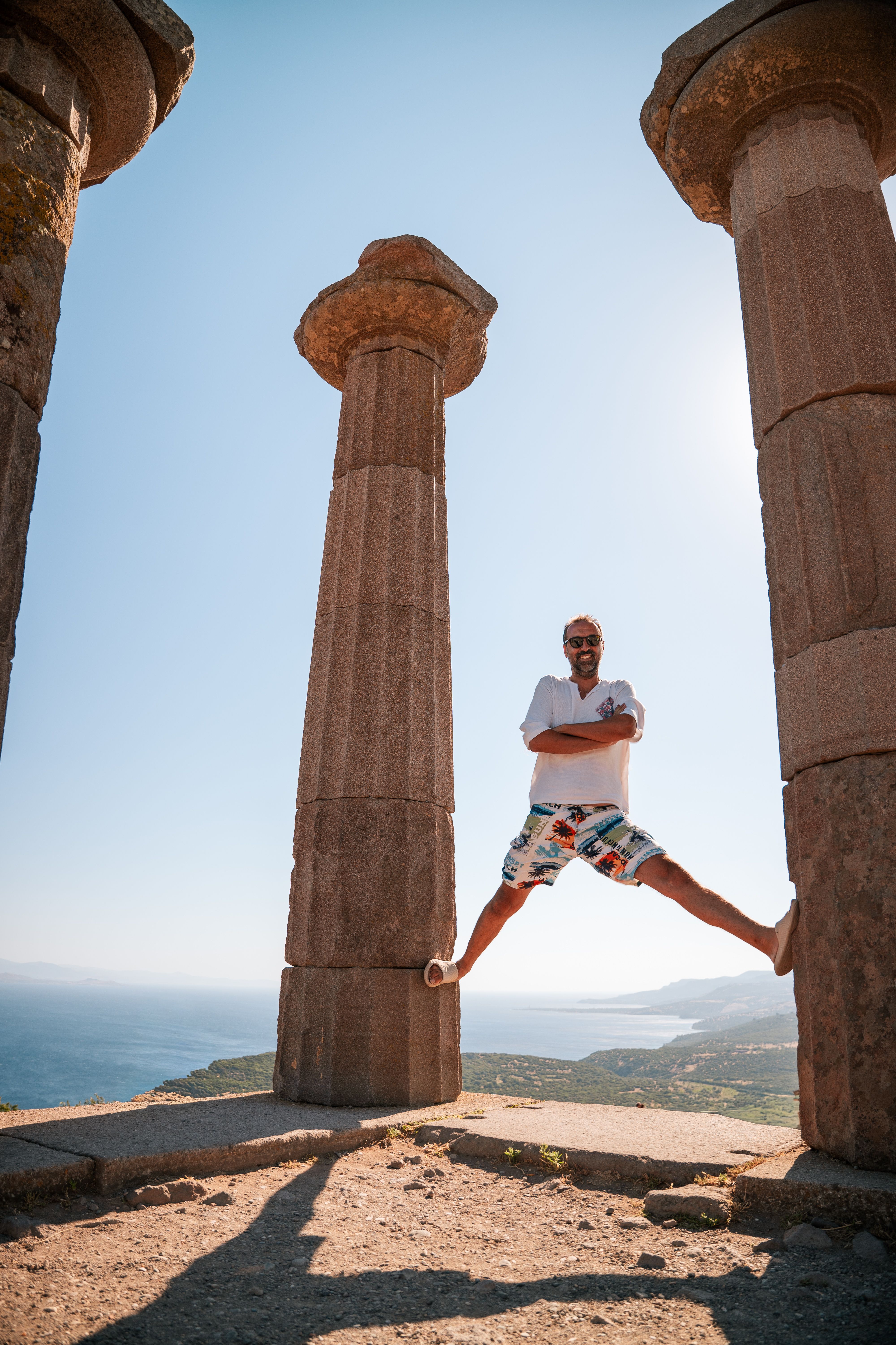A man energetically jumps between ancient Greek columns with a scenic coastal backdrop.
