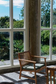 A sunlit room with a wooden chair overlooking a lush garden through large windows.