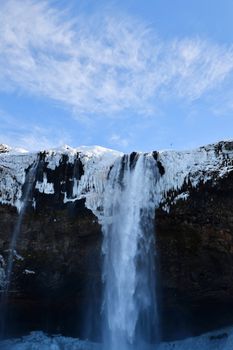 Icelandic waterfall cascading over snowy cliffs under a clear blue sky.