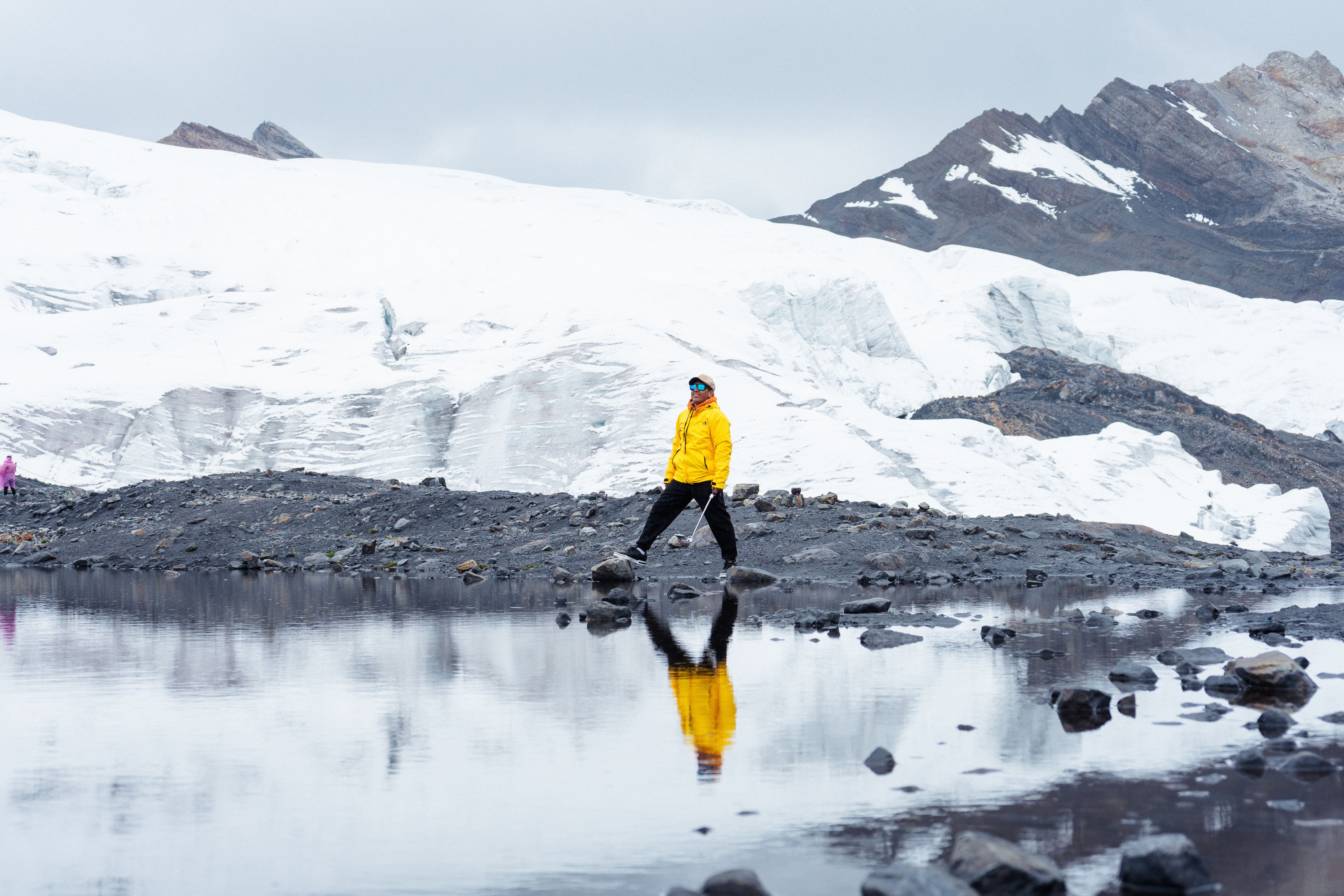 Amazing Private Tour from Quito: Hummingbirds, Waterfalls and Half the World Review 2026 - Person in yellow jacket walks by a reflective glacier pond, showcasing a serene and cold environment.