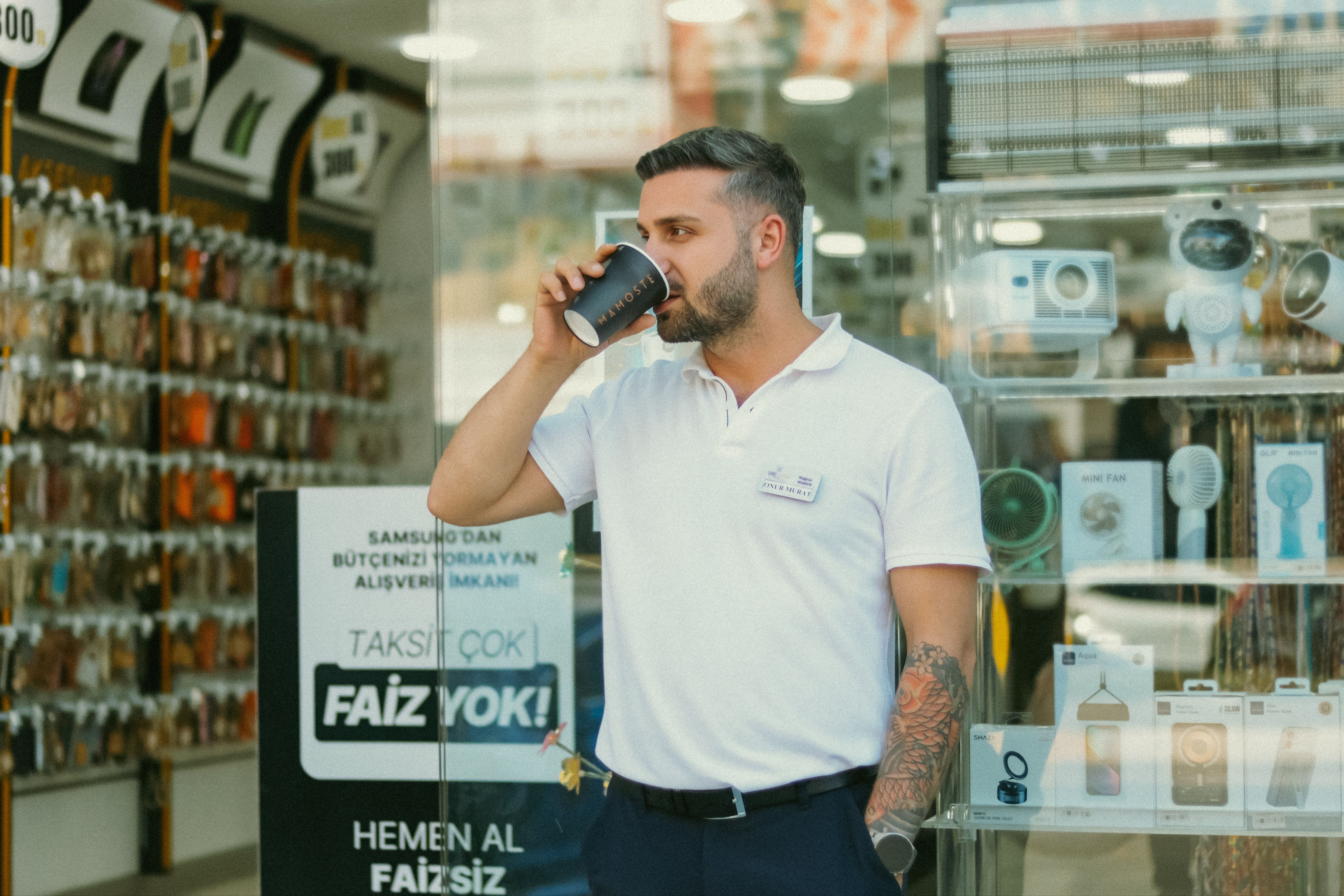 Samsung Unpacked 2024: Yeni Galaxy Ekosistemi Geliyor! - Young man sips coffee while standing in front of an electronics shop, surrounded by gadgets.
