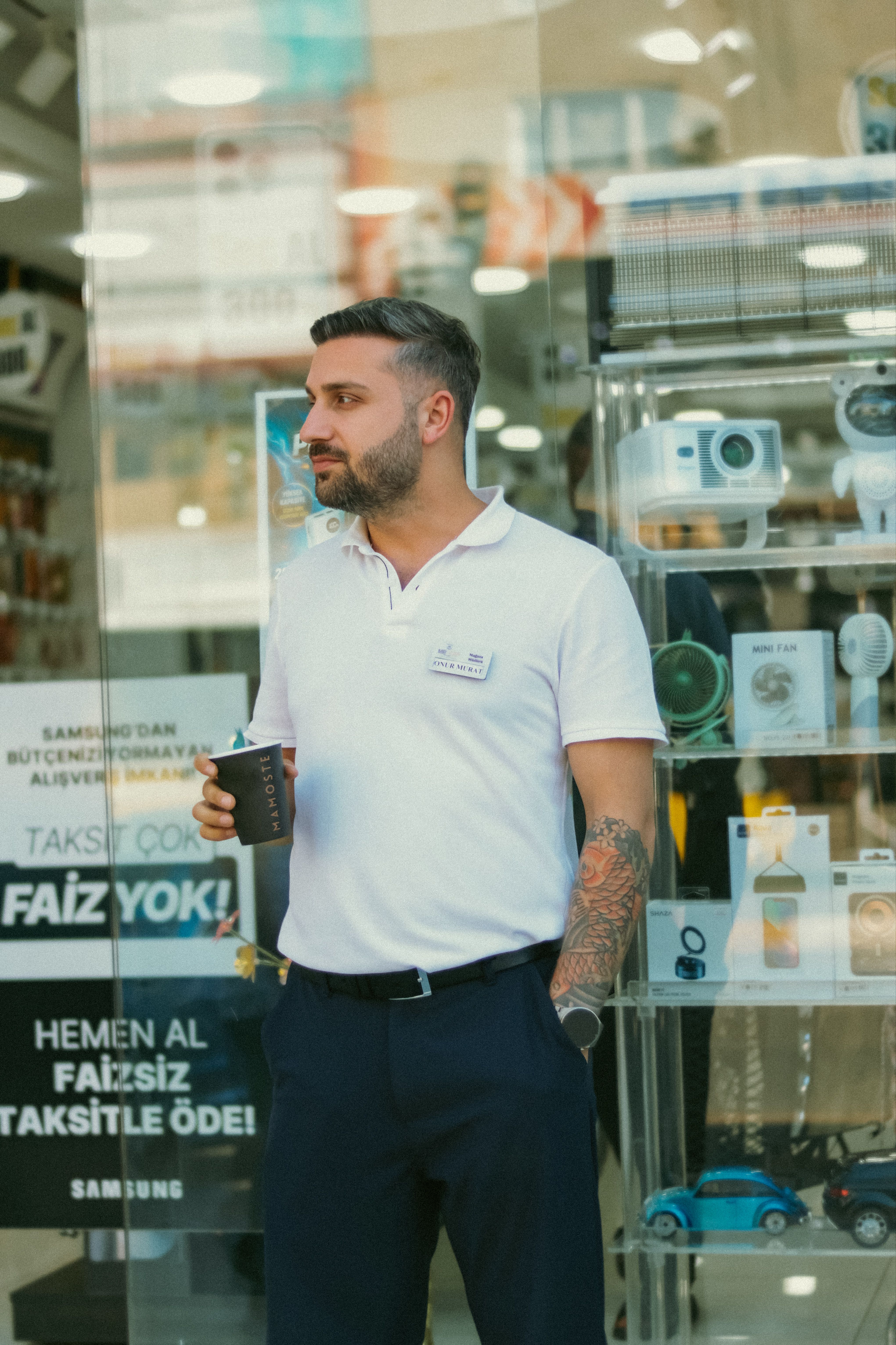 Casually dressed man with tattoos, holding coffee in front of a shop window.