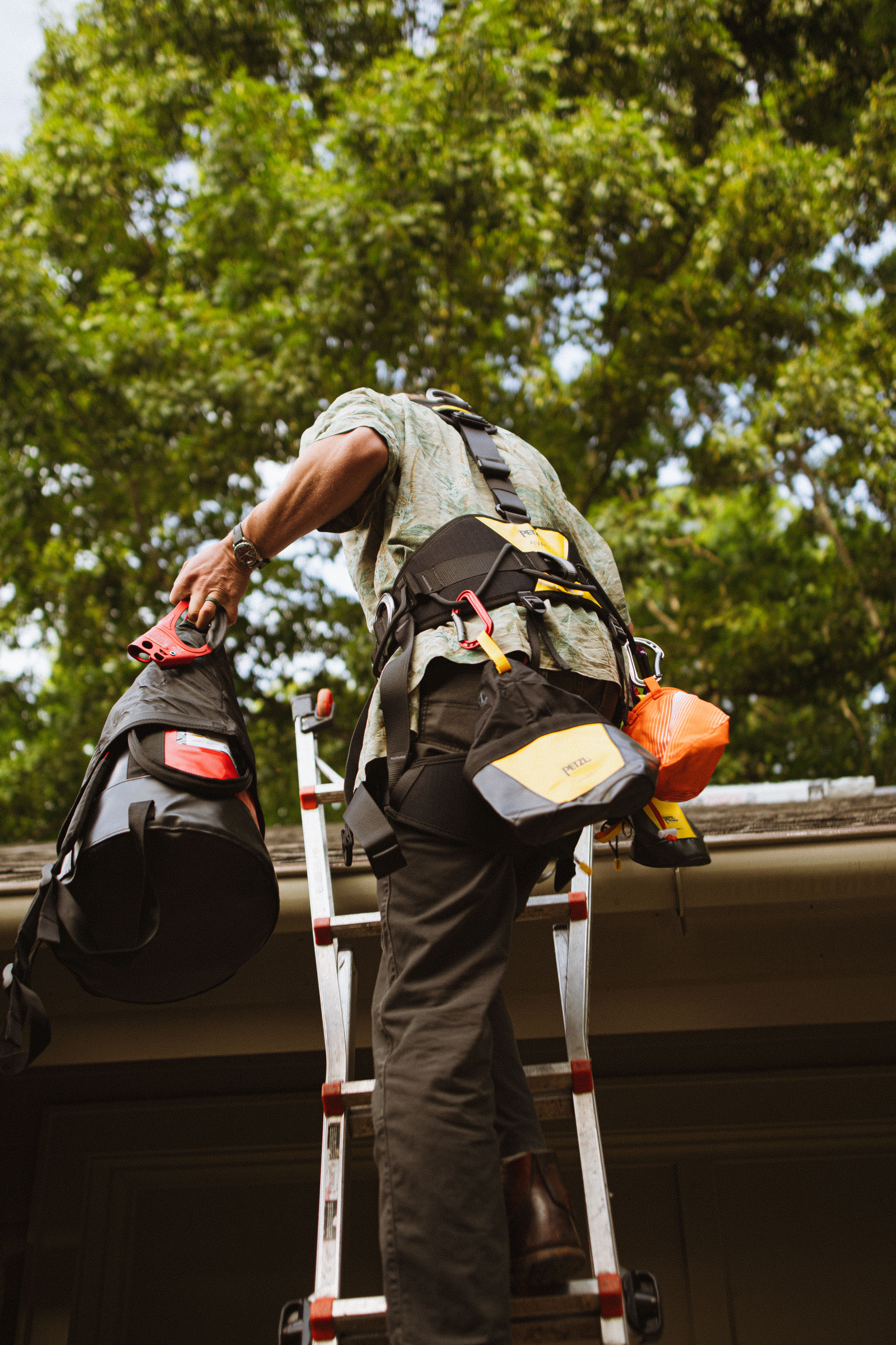 A worker in a harness with tool bags climbing a ladder toward a roofline, the kind of gear real height work calls for