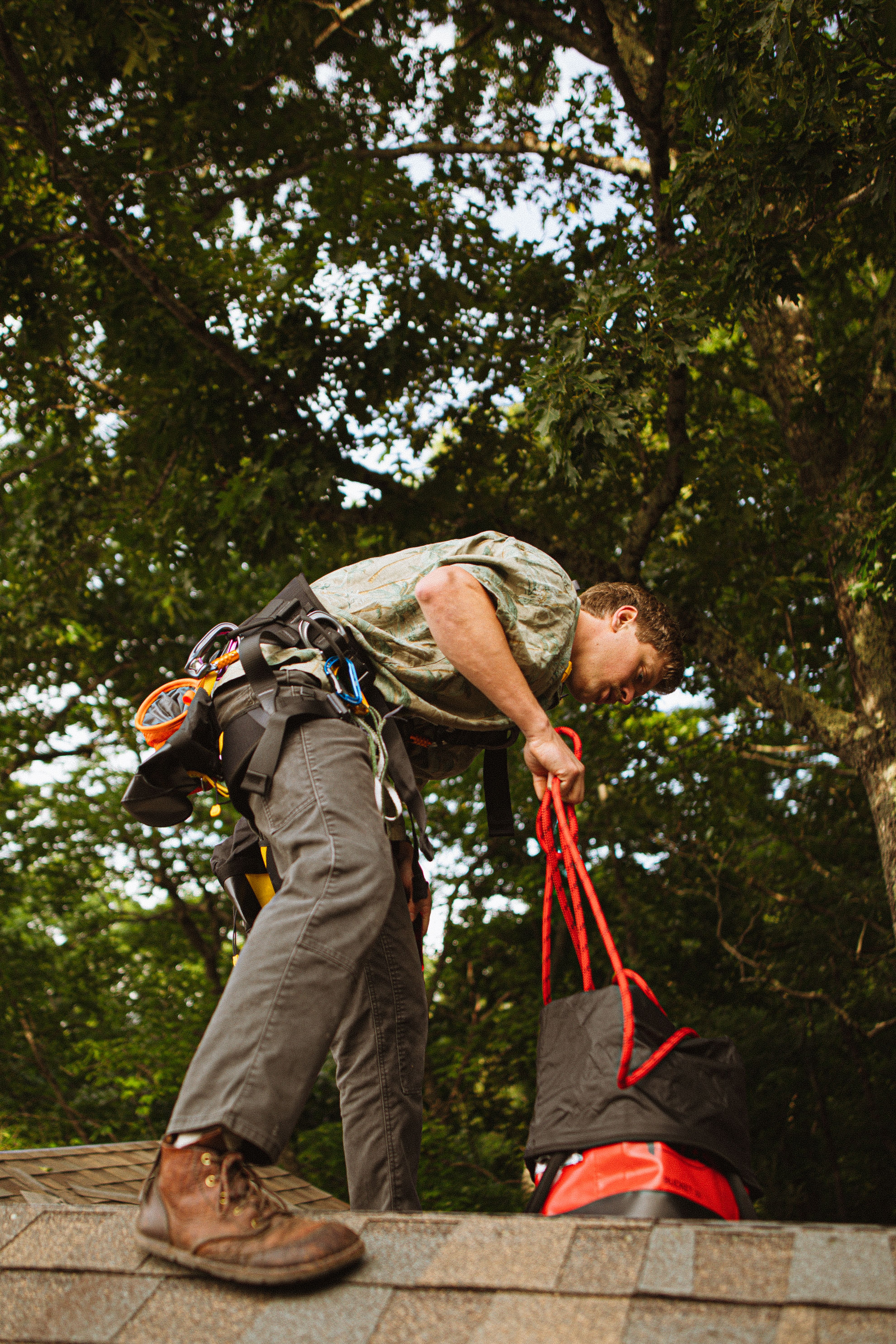 Construction worker securing equipment on a roof amid lush trees.