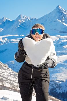 Smiling woman enjoying winter with heart-shaped snow in scenic mountain backdrop.