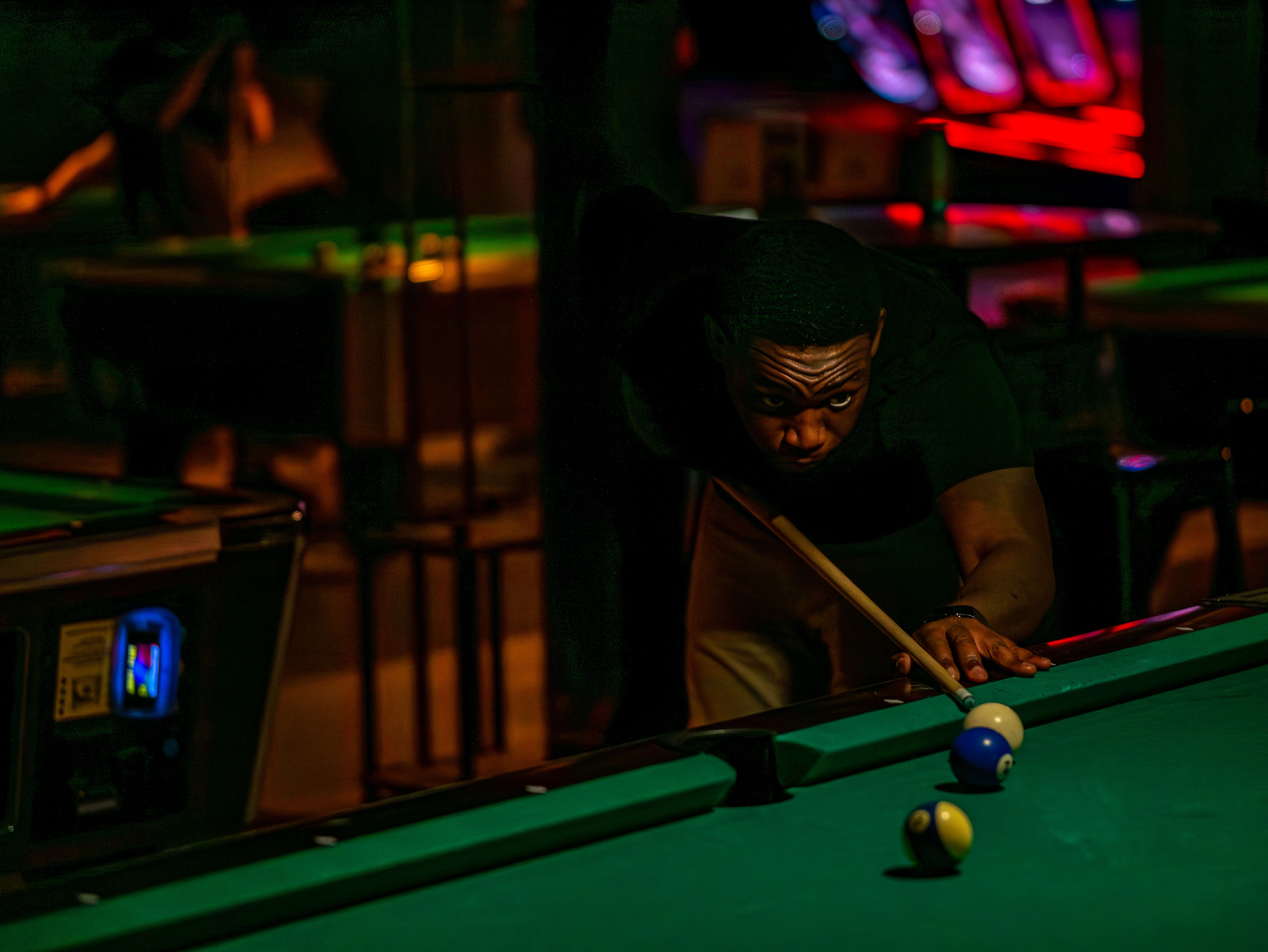 An adult man intensely focuses on a shot during a competitive game of pool in a dimly lit billiards hall.