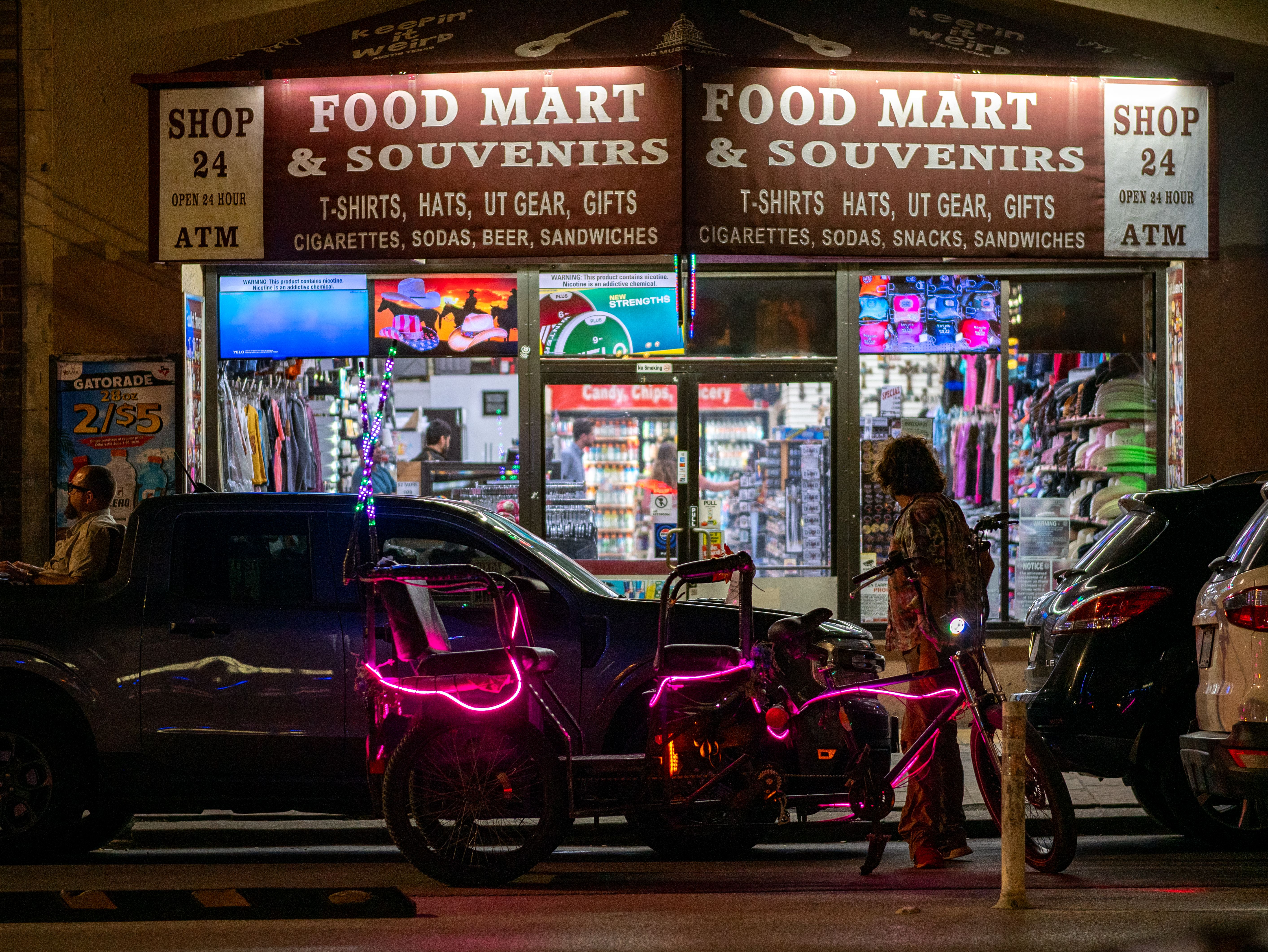 Neon-lit bike parked outside a bustling city food mart at night with colorful window displays.
