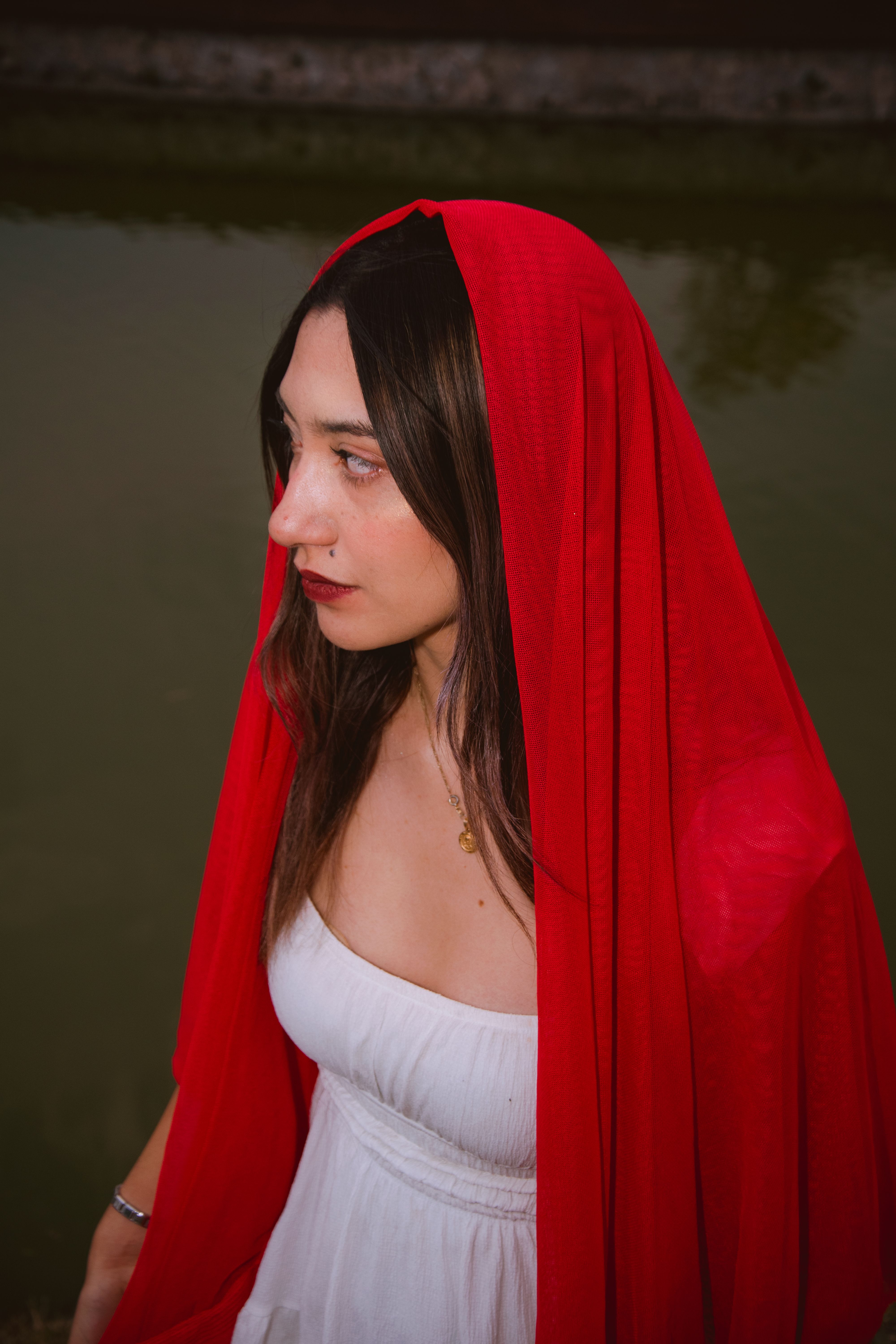 A woman in a red veil stands by the water, captured in serene contemplation.