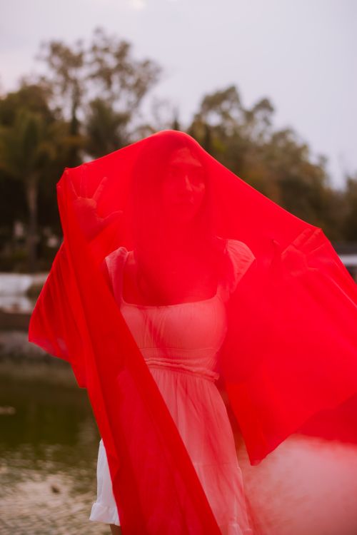 Free A woman in a white dress is wrapped in a flowing red veil, standing outdoors near water, creating a mystical atmosphere. Stock Photo
