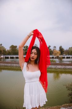 A young woman in a white dress holding a red scarf by a pond in a park.