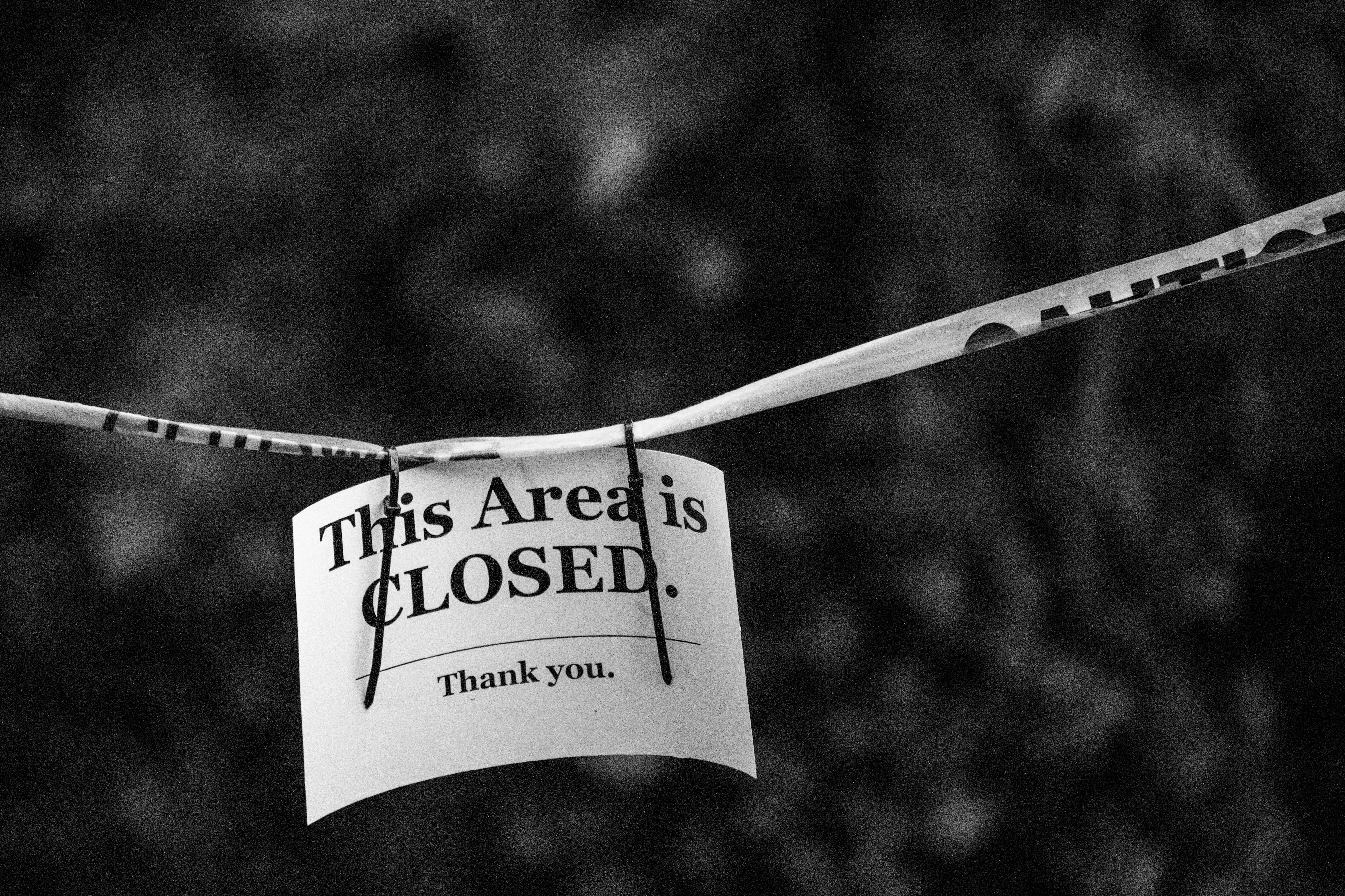 A black and white photo of a sign reading 'This Area is CLOSED.' in a blurred outdoor setting.