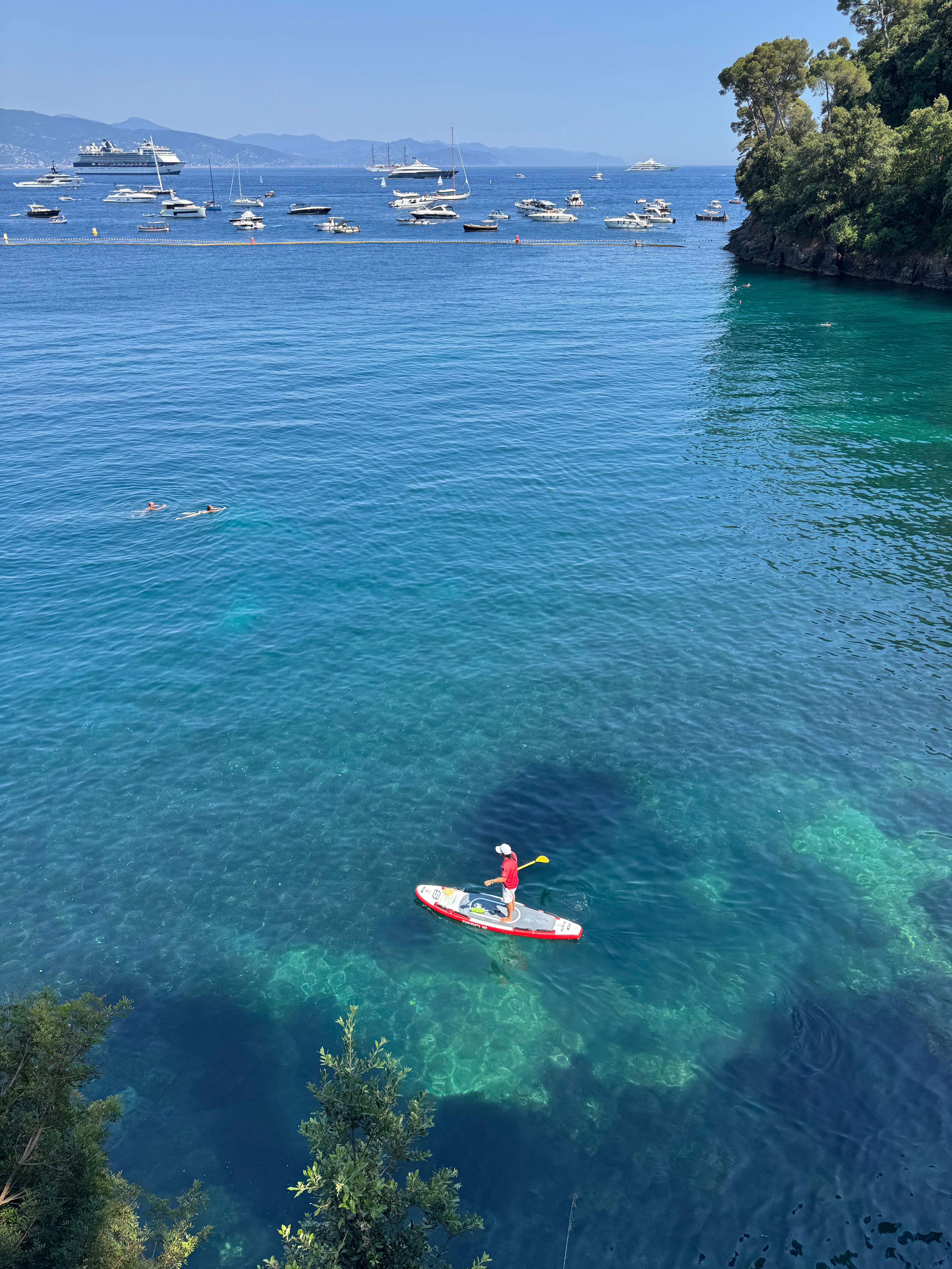A paddleboarder enjoys the crystal-clear waters of Portofino, Italy, with yachts in the distance.