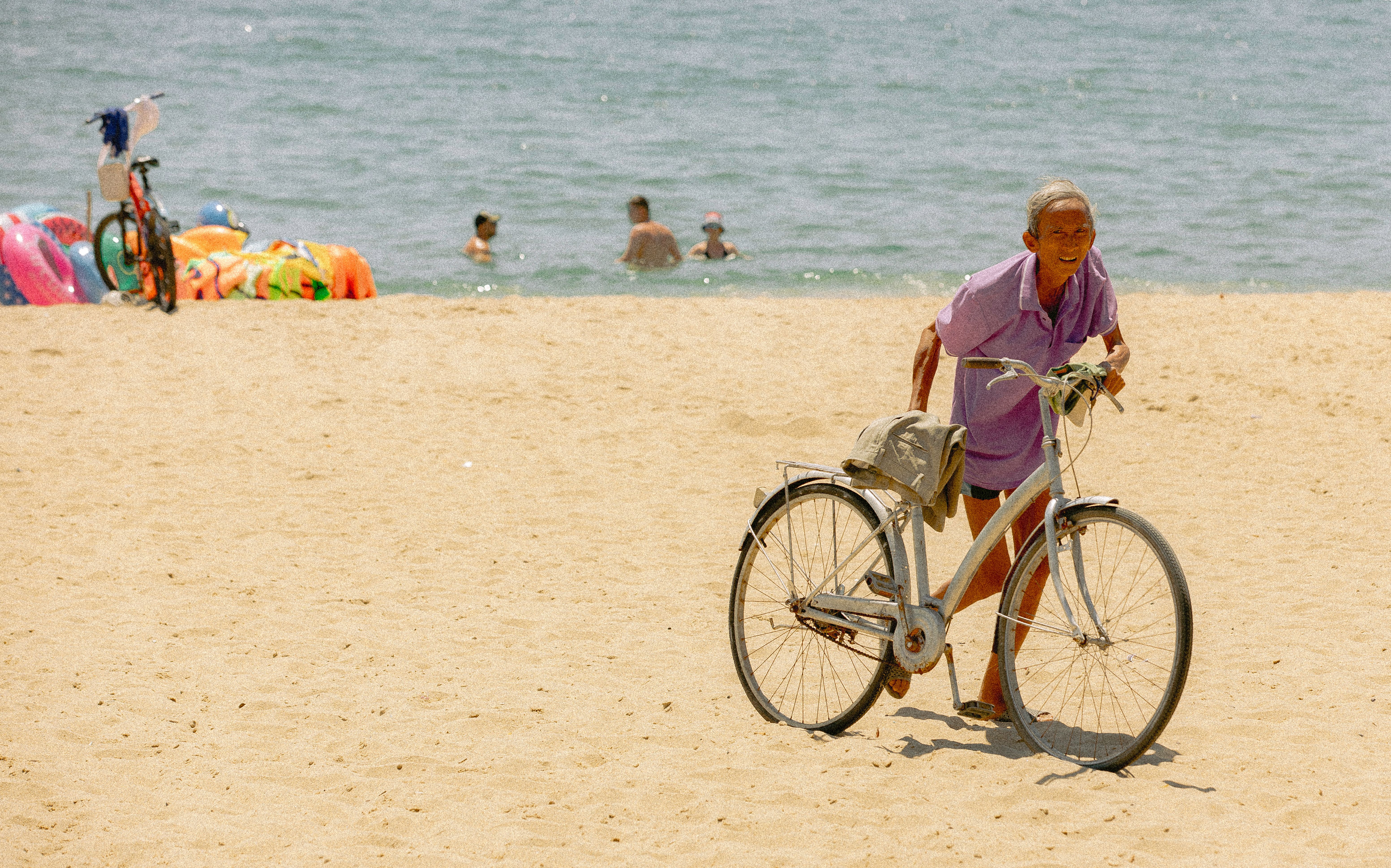 An elderly man pushes a bicycle along a sunny, sandy beach with people swimming in the background.