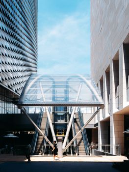 A transparent escalator tunnel between two modern buildings under a bright daytime sky.