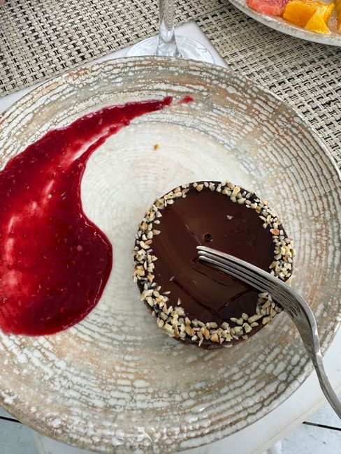 A small pitcher of glossy red raspberry coulis being poured over a white plate, with a fine mesh strainer and fresh raspberries beside it