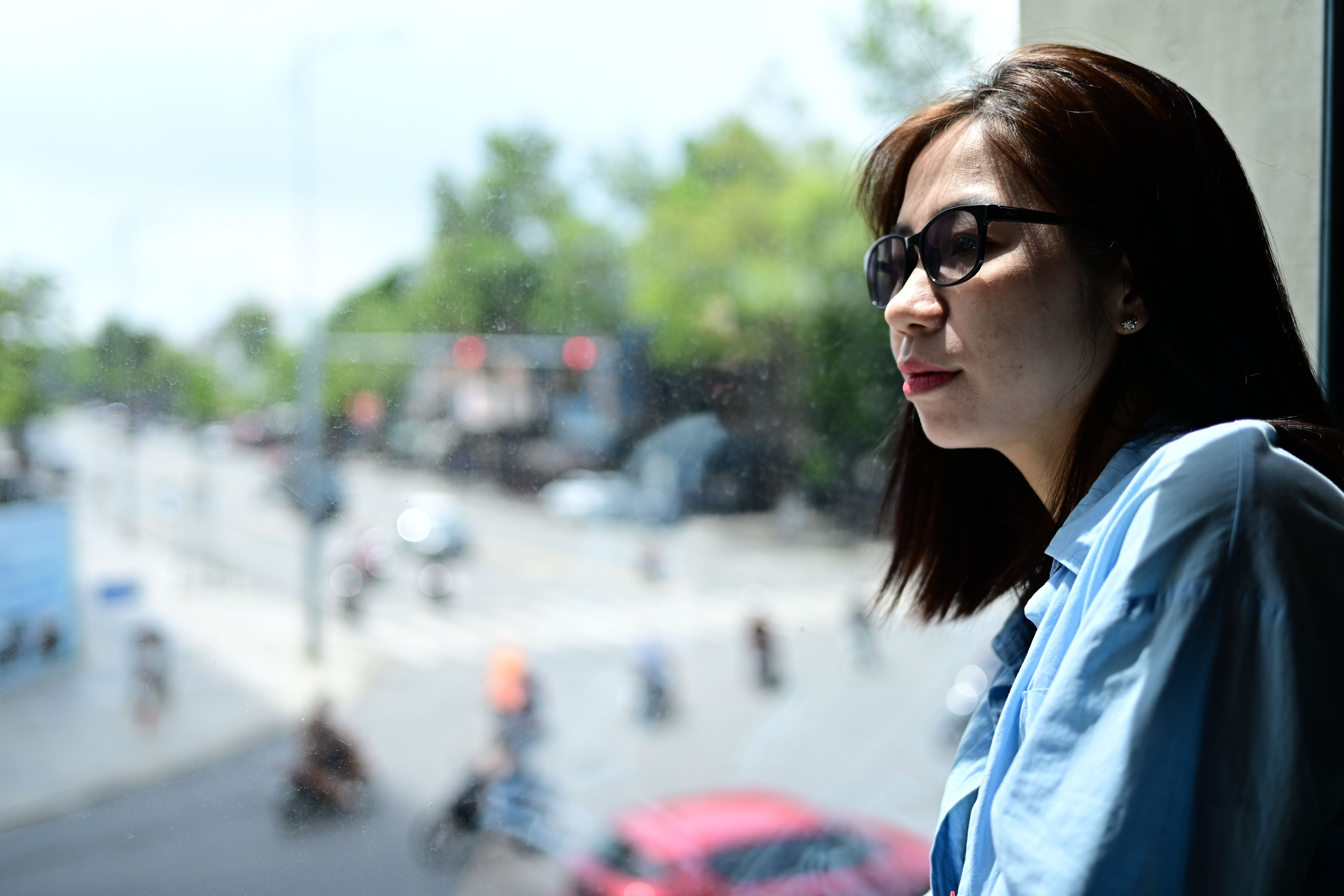 A thoughtful woman with glasses looks out a window, capturing a blurred urban scene.