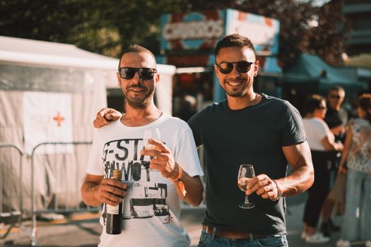 Two friends enjoying a sunny festival in Switzerland with wine and sunglasses.