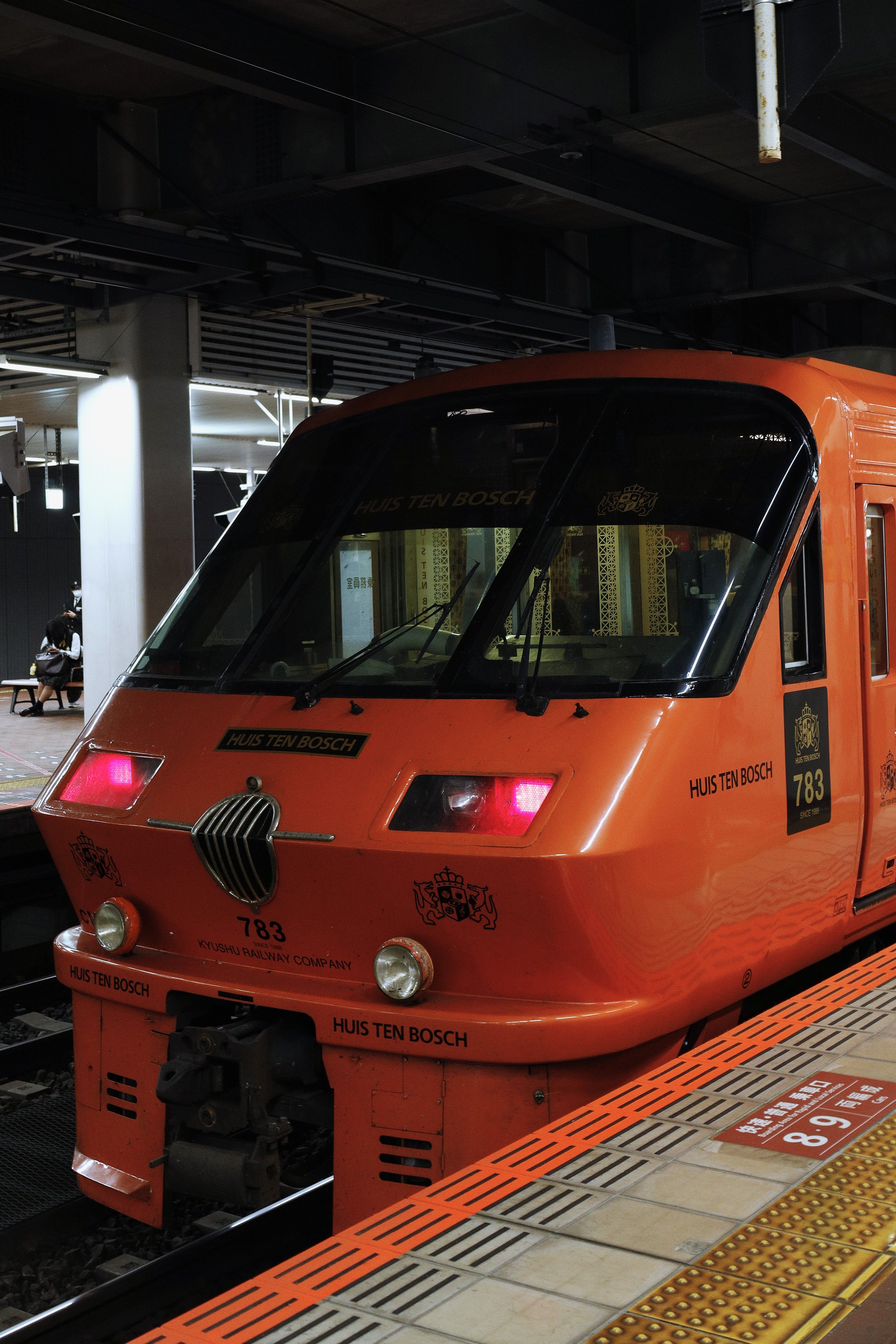 Orange Huis Ten Bosch train at Fukuoka station platform in Japan, showcasing modern rail travel.