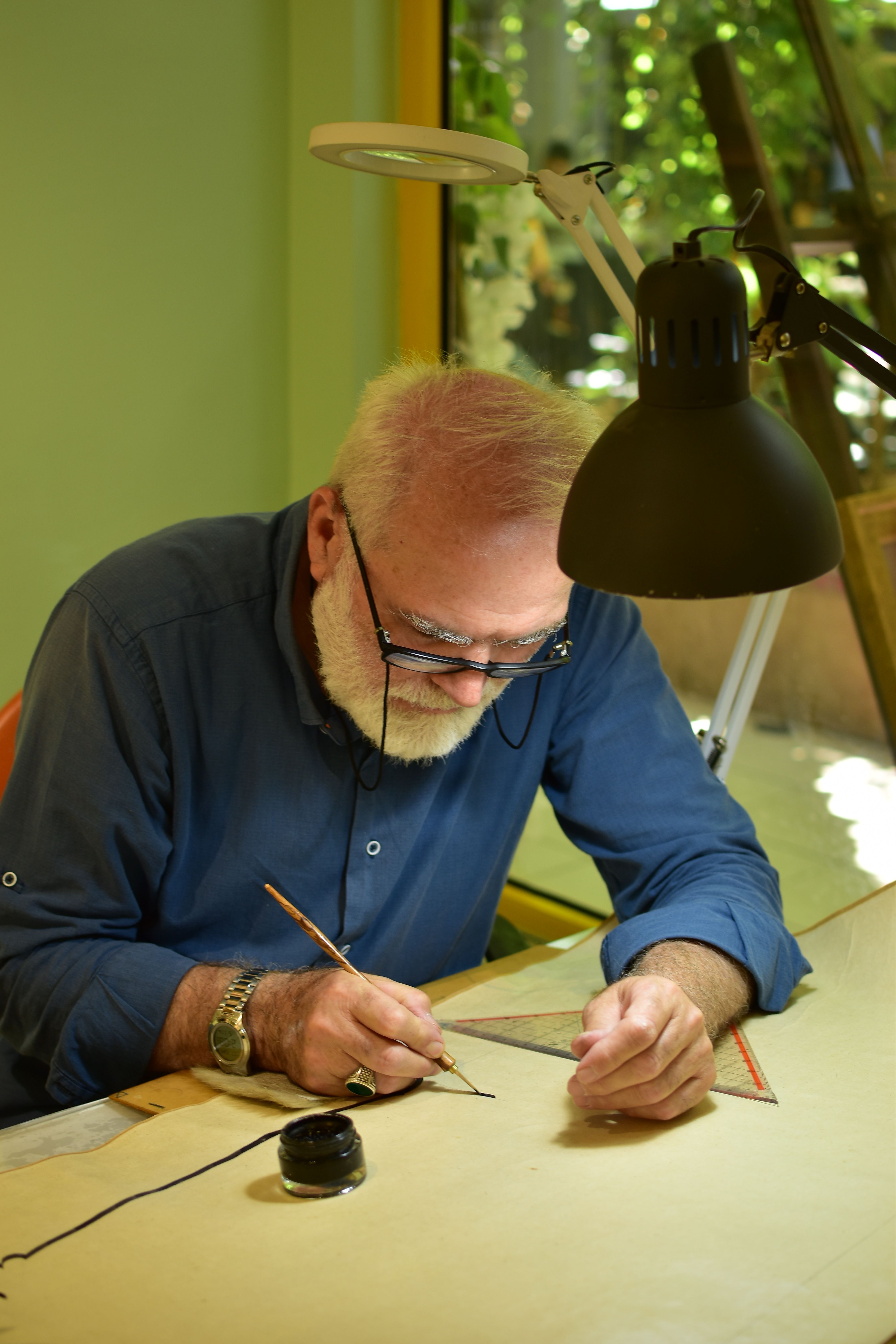 An elderly man concentrating on an intricate ink drawing in a well-lit studio setting.