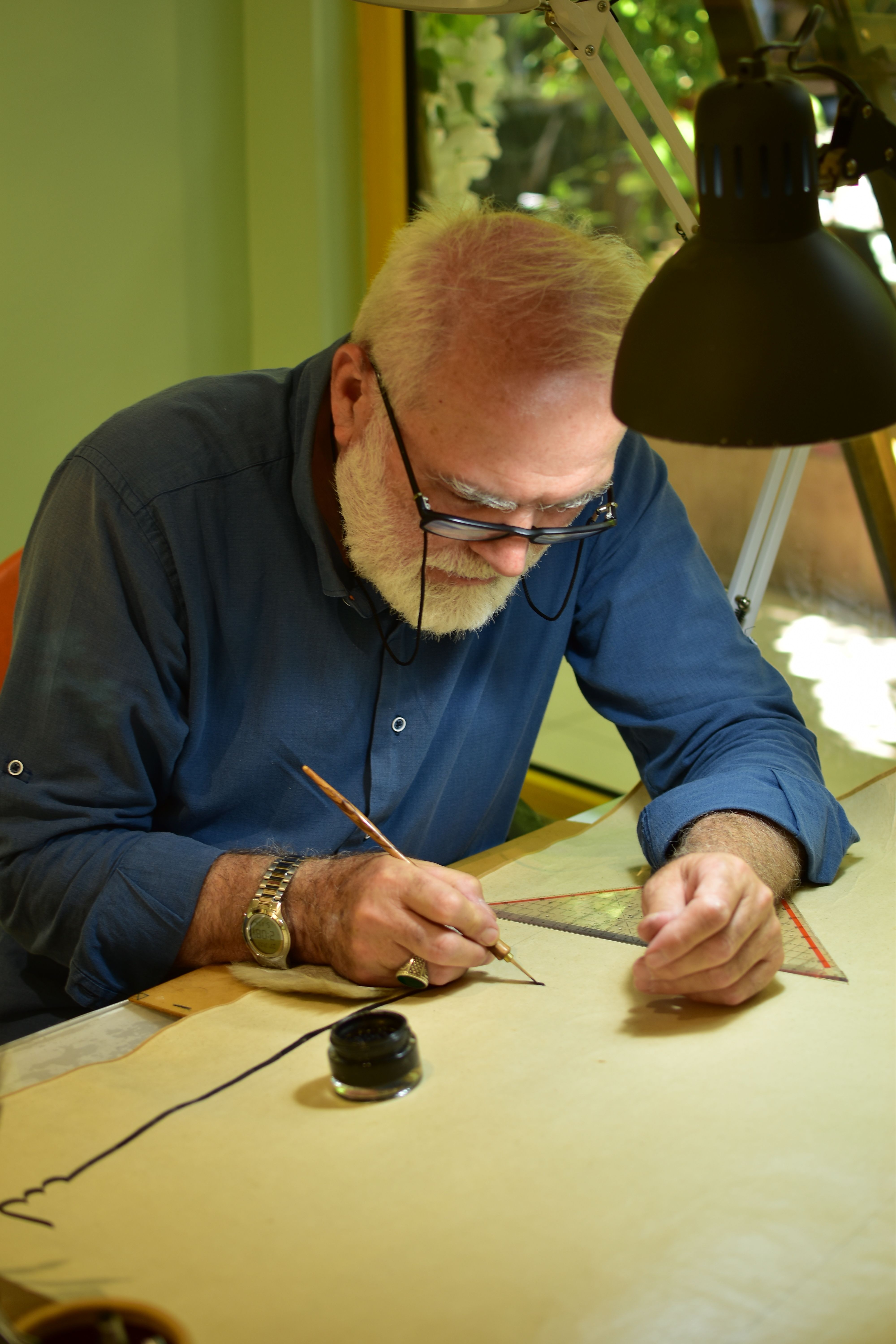 Senior male artist focused on drawing with an ink pen at a well-lit desk indoors.