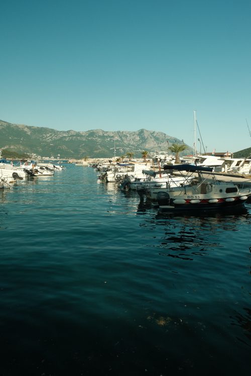 Free Idyllic Budva Marina with boats docked, surrounded by picturesque mountains and clear blue sky. Stock Photo