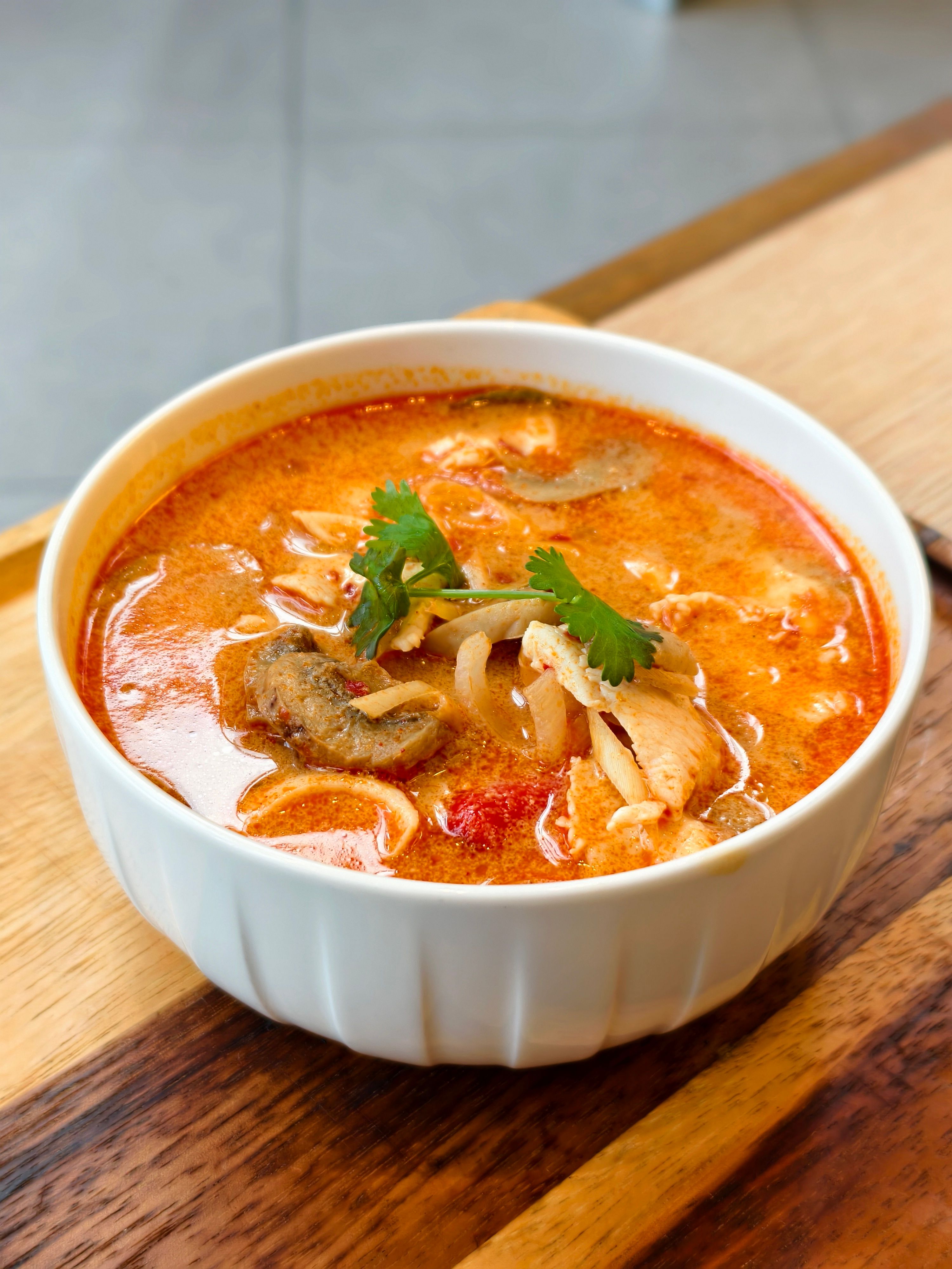 Close-up of a spicy Tom Yum soup with chicken, mushrooms, and cilantro in a white bowl.