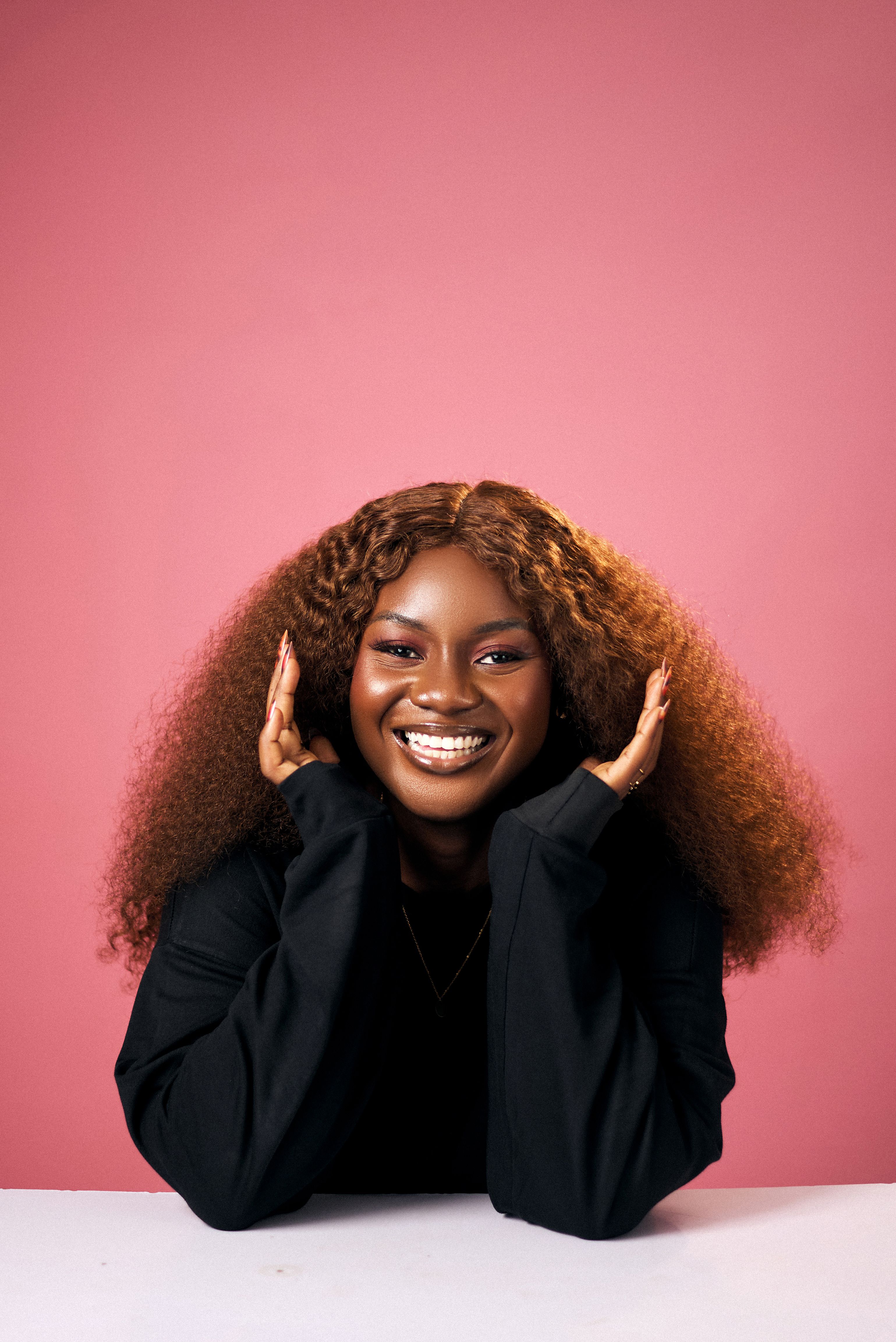Portrait of a young woman with curly hair smiling and posing over a pink background.