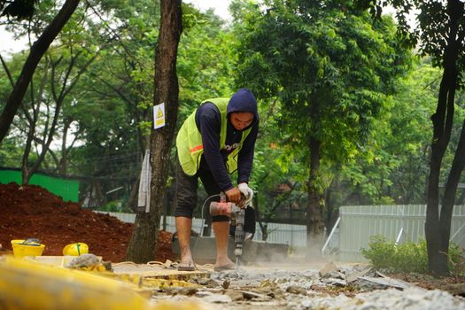 A construction worker in casual attire operates a drill on a site surrounded by greenery.