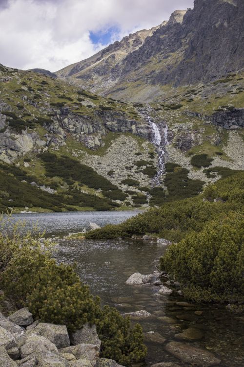 Free Majestic waterfall cascading down a rugged mountain in an alpine setting under a cloudy sky. Stock Photo