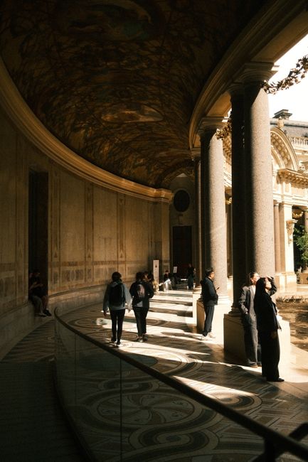 A grand hallway with ornate ceiling and people illuminated by sunlight.