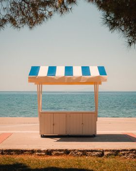 A blue and white striped vendor stand facing a serene sea under a clear sky, perfect for summer vibes.