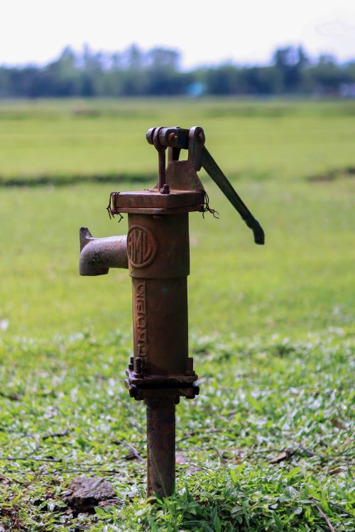 Free Rustic hand water pump surrounded by lush green field, representing traditional irrigation. Stock Photo