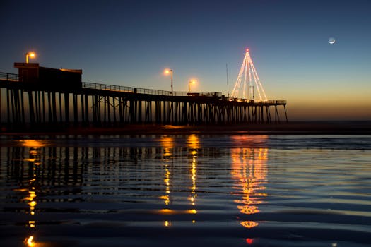 Illuminated Pismo Beach Pier at twilight with festive lights reflecting on the ocean and crescent moon in the sky.