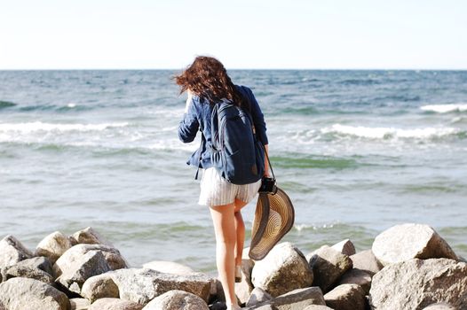 A young woman with a backpack walks on a rocky beach, enjoying a sunny summer day by the sea.