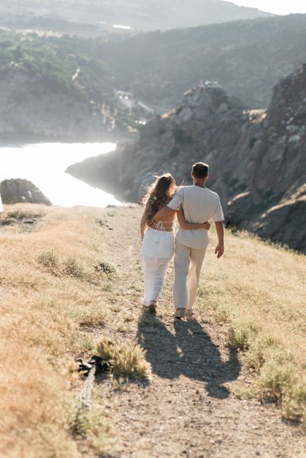Couple walking arm in arm along a sunny mountain path with a scenic view.